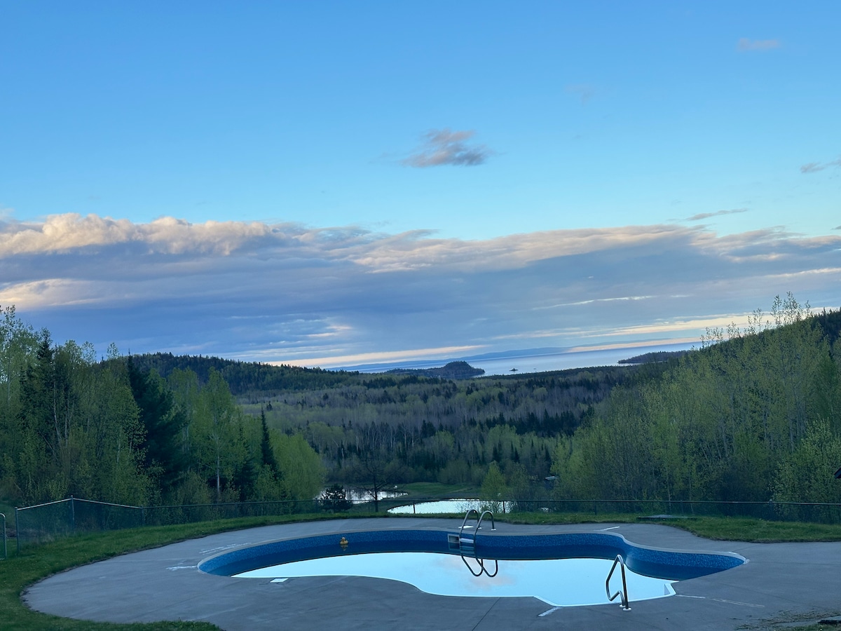 A serene swimming pool is positioned on a patio, reflecting the blue sky above. Lush greenery surrounds the area, while distant views of the lake and hills create a peaceful backdrop. The landscape reveals soft, rolling terrain under a cloud-dotted sky.