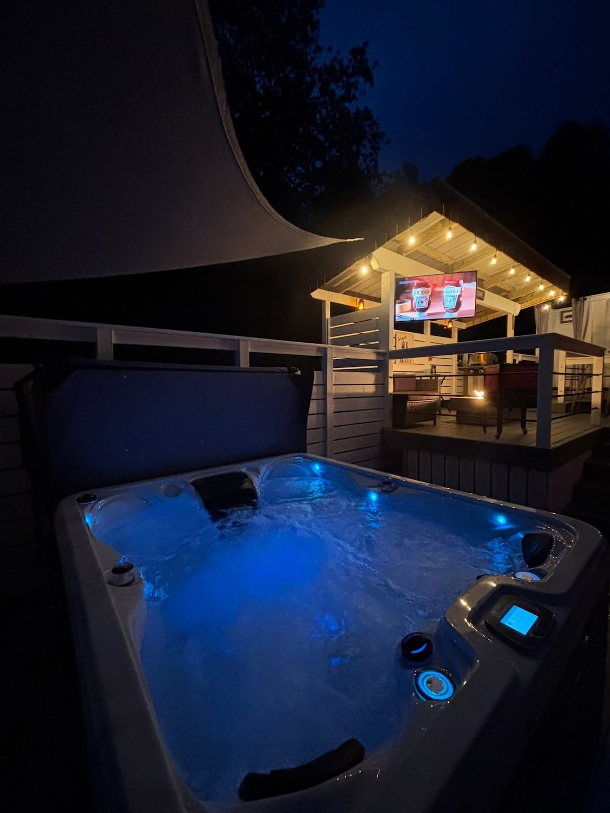 An outdoor hot tub is illuminated with soft blue lighting, inviting relaxation. A nearby covered seating area is equipped with string lights, providing a cozy atmosphere at night. A television is mounted above the seating area, enhancing entertainment options under the stars.