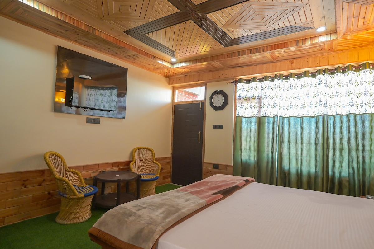 A spacious room features wooden paneling and a coffered ceiling, enhancing the sense of warmth. A large bed is positioned in front of a flat-screen TV. Two wicker chairs surround a small table, with a door leading outside and large windows dressed in patterned curtains.