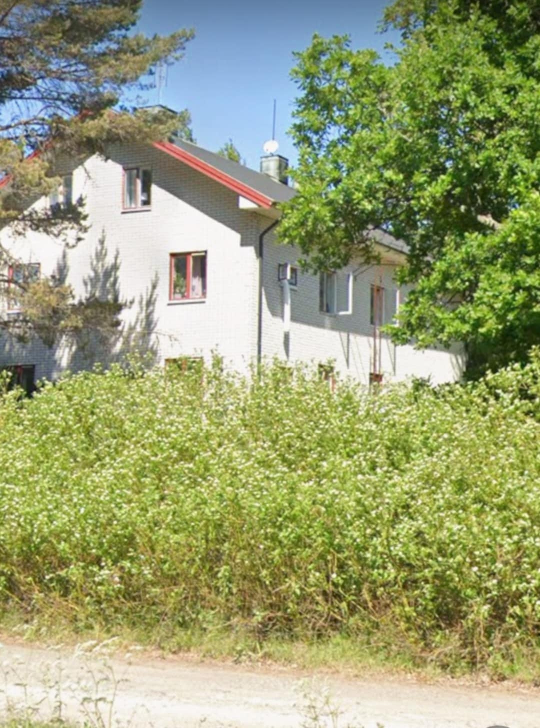The exterior of a two-story building is surrounded by lush greenery and wildflowers. The white brick structure features multiple windows and a red roof. Tall trees provide shade, creating a serene and natural setting that enhances the peaceful environment.