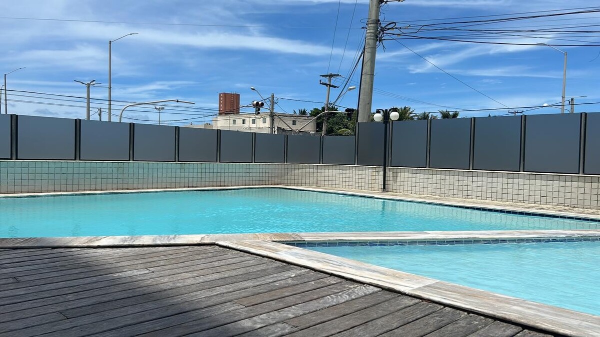 A private swimming pool is displayed, featuring clear blue water and surrounded by a wooden deck. Tall privacy screens line the perimeter, creating an enclosed space. Sunlight reflects on the water's surface, enhancing the overall brightness of the area.