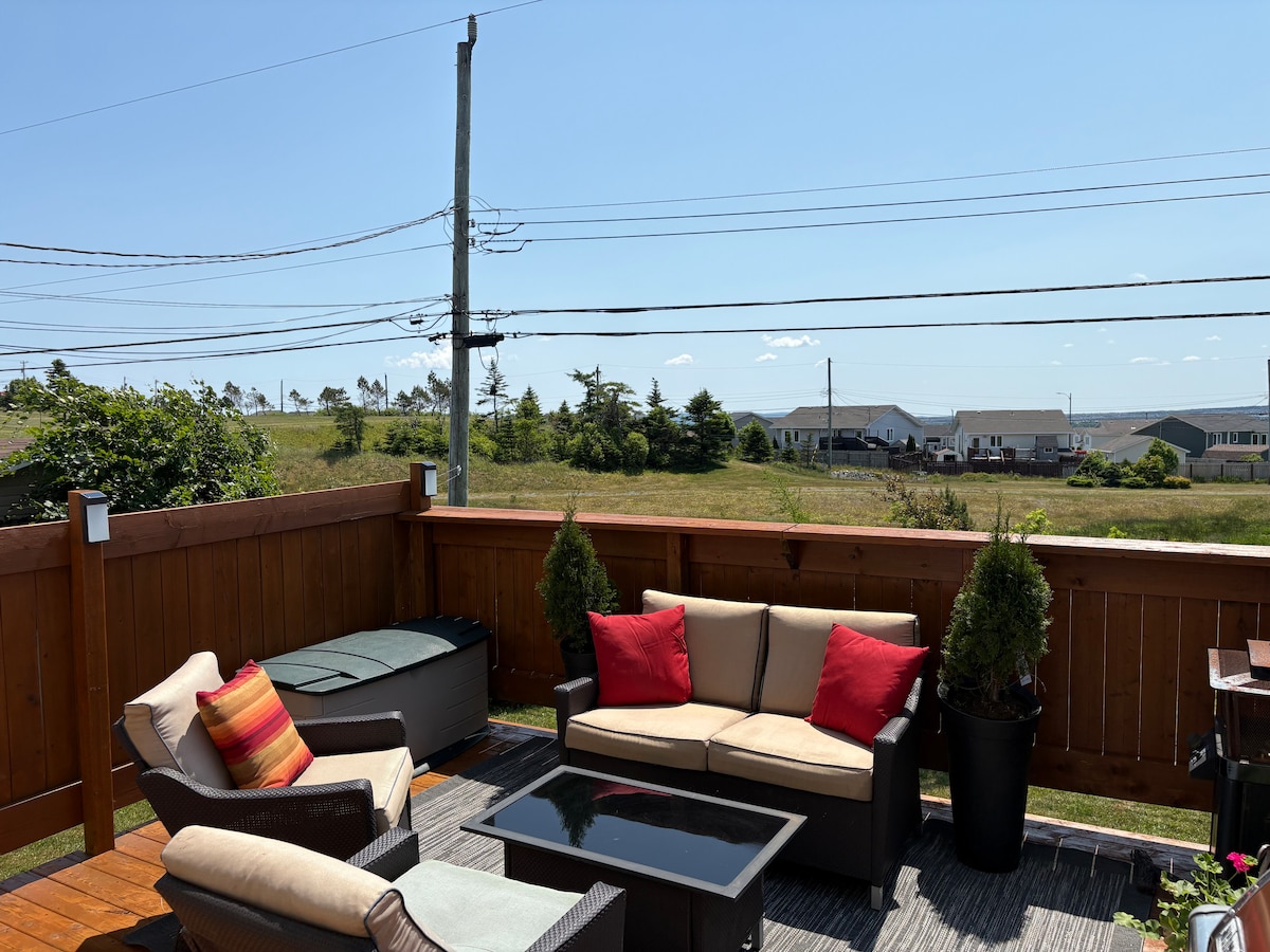 An outdoor deck is presented with comfortable seating, including a sofa adorned with red accent pillows, surrounding a glass-top table. Lush greenery and views of a serene landscape are visible, with distant houses under a clear blue sky.