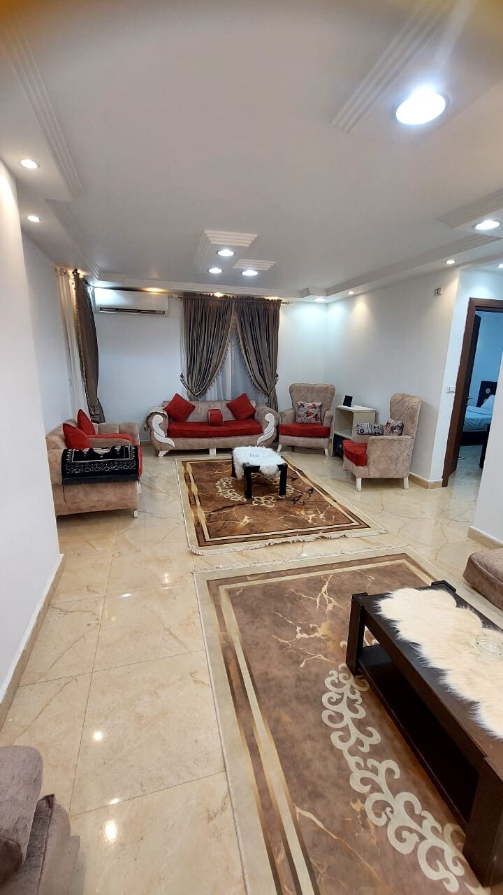 A welcoming living area is presented with soft, neutral tones and pops of red accents from cushions. Comfortable seating is arranged around a decorative rug, with a simple coffee table positioned centrally. Ambient lighting enhances the inviting ambiance, contributing to a warm, relaxed environment.