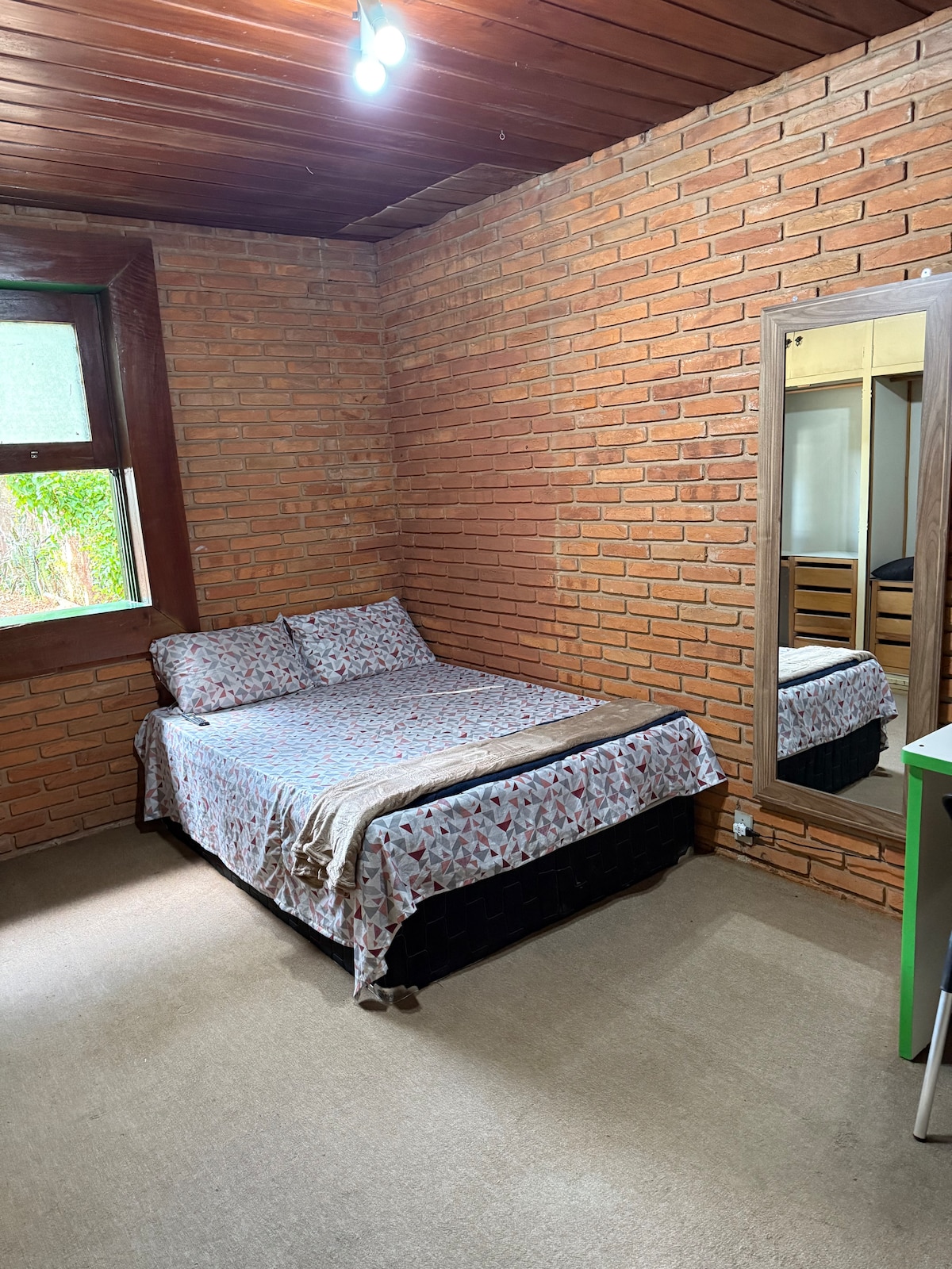 A spacious guest room features a large bed adorned with a patterned blanket. Exposed brick walls add warmth, while a full-length mirror reflects the inviting space. Natural light filters in through the window, highlighting a simple desk and chair in the corner.