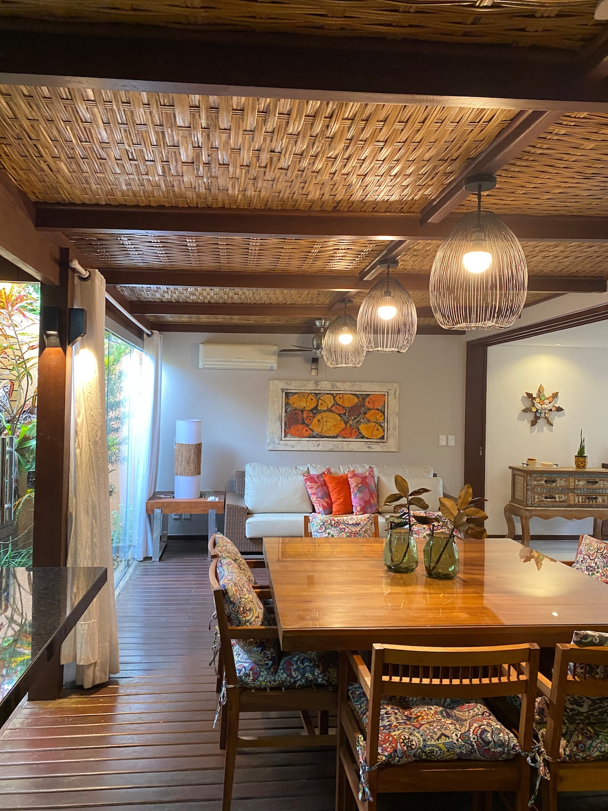 An open living and dining area features a wooden dining table surrounded by floral-patterned chairs, inviting seating, and decorative plants. Natural light filters through large glass doors, illuminating the space. Light fixtures with woven designs hang from a textured ceiling, enhancing the cozy ambiance.