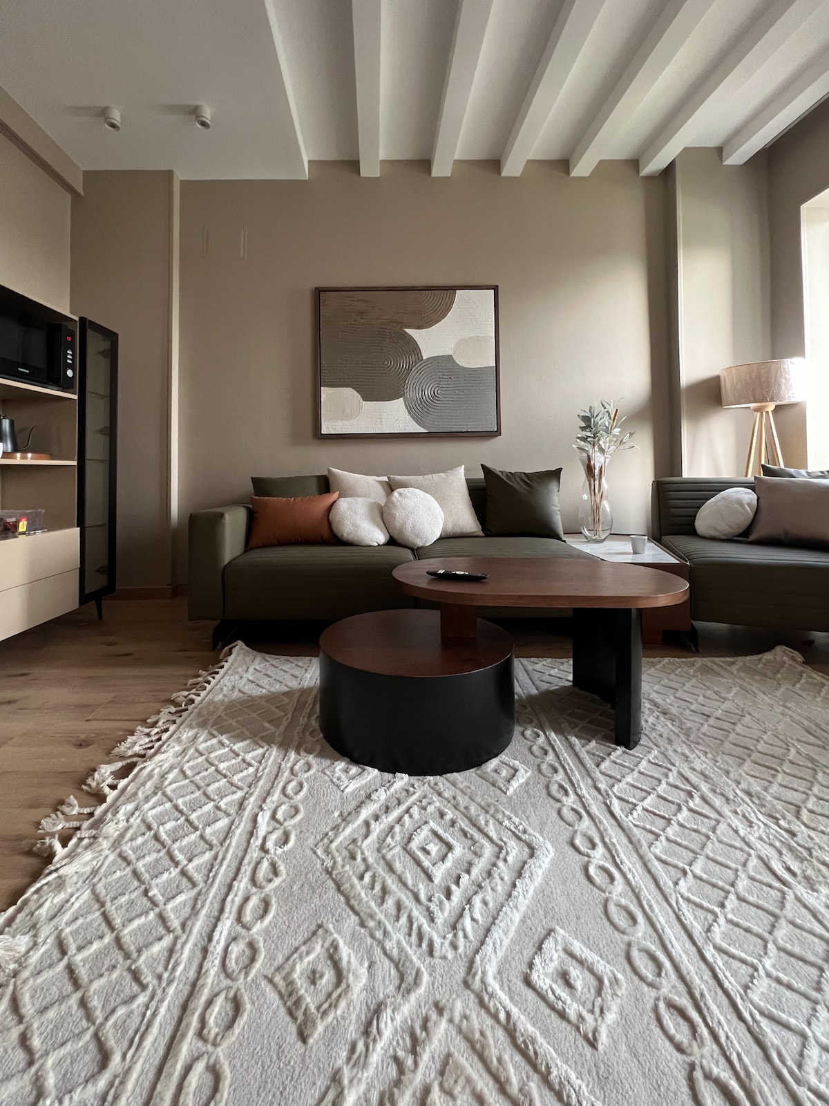 A comfortable living area features a modern sectional sofa adorned with soft cushions in earthy tones. A round coffee table rests on a textured area rug, and a stylish floor lamp stands beside the window, creating a warm and inviting ambiance.