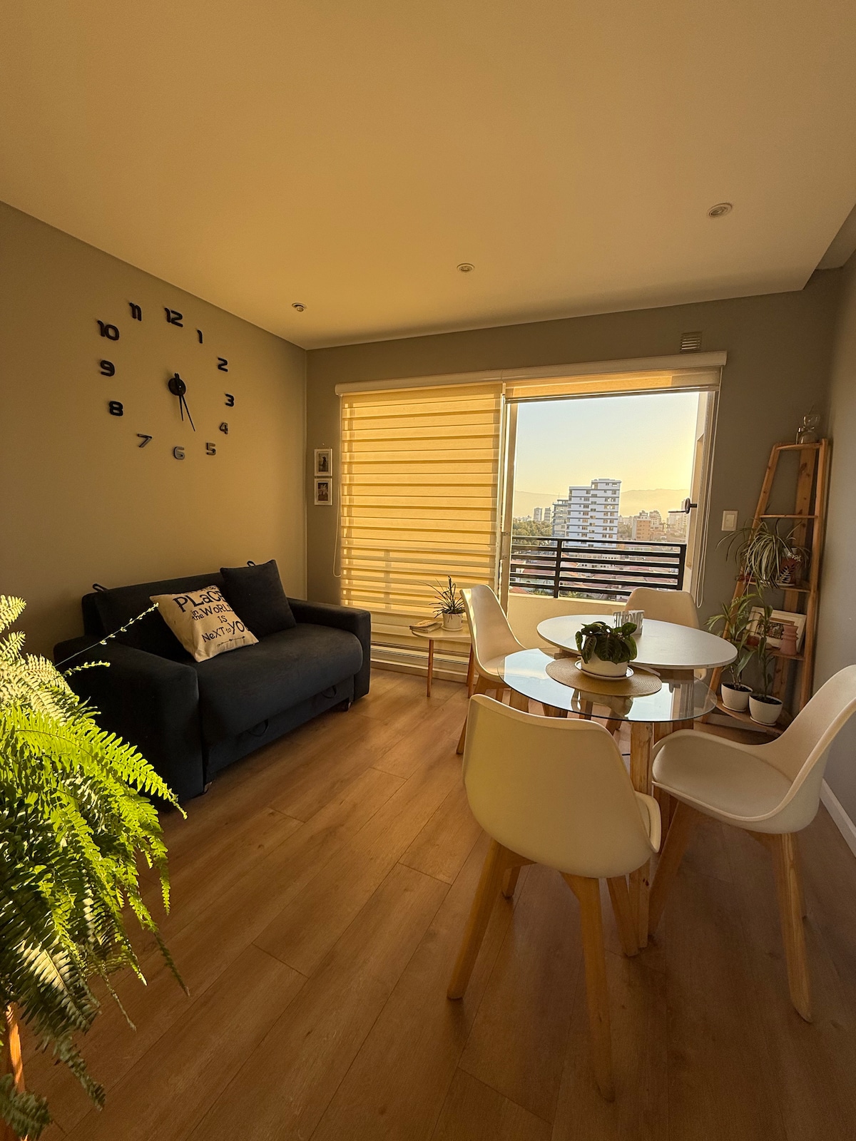 A bright living area is highlighted by a large window with layered shades, offering a view of the city skyline. A round dining table is surrounded by white chairs, while a dark sofa and wall clock add practical touches to the modern design.