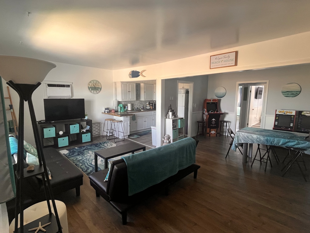 An open living area features a combination of seating including futons and chairs, alongside a coffee table. A kitchen area is visible with modern appliances and a bar-style table. Natural light fills the space, enhancing the decor with beach-themed accents.
