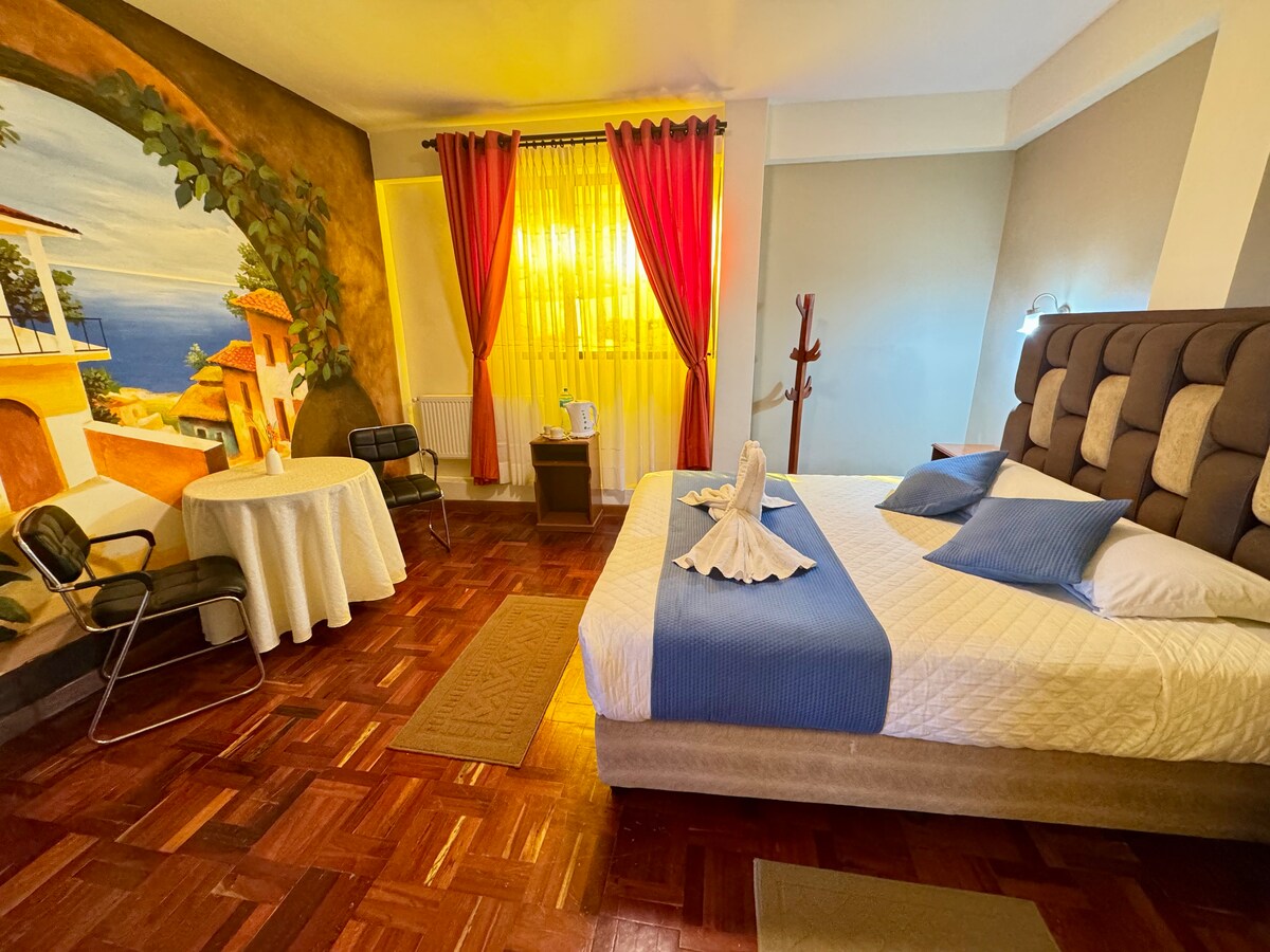 A spacious room features a comfortable queen-size bed, complemented by decorative pillows. A round table with chairs is present for work or leisure. Large windows allow natural light to illuminate the hand-painted coastal mural, while curtains provide privacy.