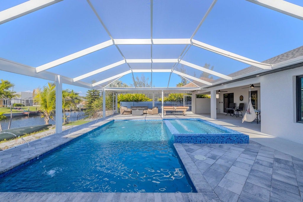 A spacious outdoor area features a large swimming pool and a hot tub, all under a screened enclosure. Sun loungers are placed around the pool, and views of the canal are visible beyond the landscaped garden. The area is bathed in natural sunlight, providing an inviting atmosphere.
