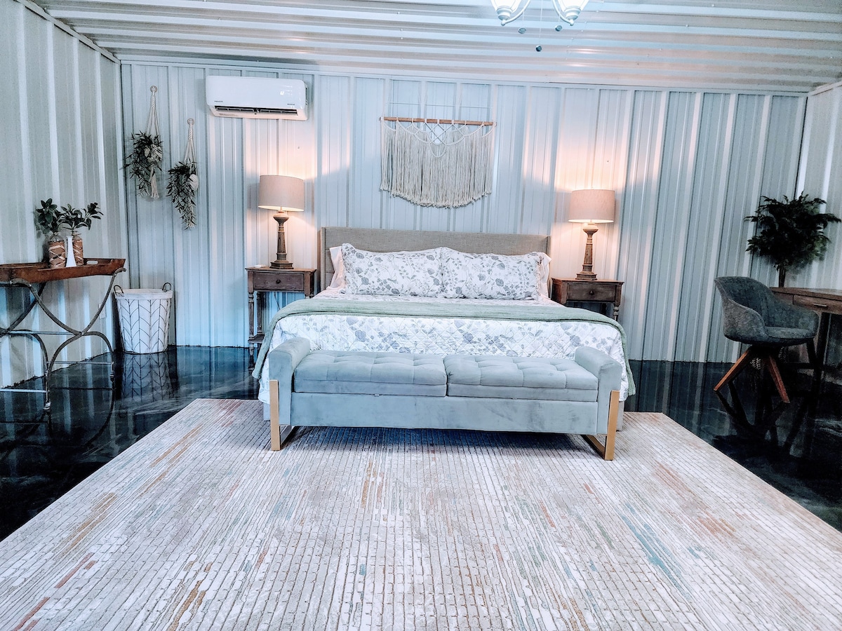 The sleeping area showcases a spacious bed with decorative pillows, positioned against a textured wall. Two bedside tables with lamps flank the bed, while a woven wall hanging adds texture. A large area rug covers the floor, and a small desk chair is visible nearby.