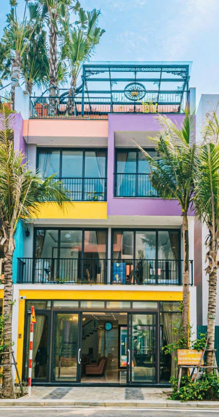 A vibrant, multi-colored three-story building is framed by tall palm trees. Large glass doors provide access to the ground floor, while balconies with black railings are seen on the upper levels. A rooftop area is visible at the top, suggesting outdoor seating.
