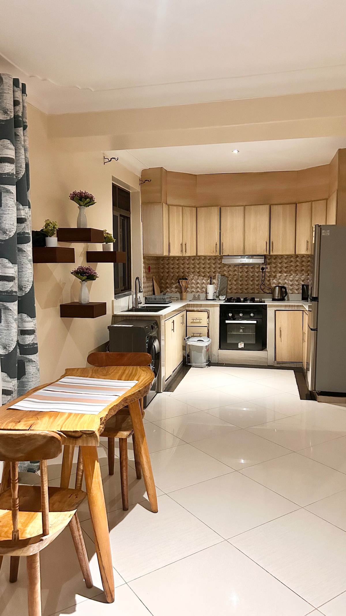 A modern kitchen features wooden cabinetry and a tiled backsplash. Stainless steel appliances are arranged along one wall. A small dining area is present with a wooden table and two chairs. Decorative plants are placed on wall-mounted shelves, enhancing the overall design.