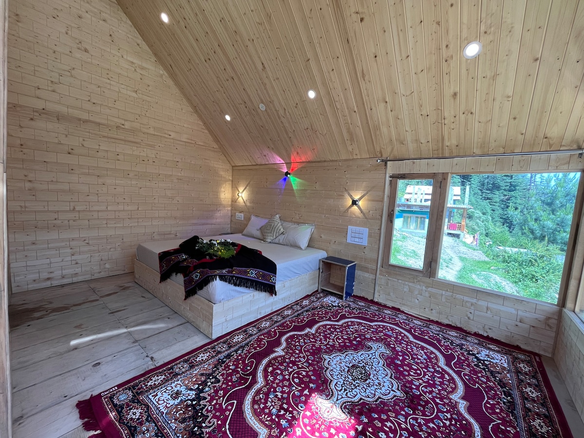 The room features a light wood interior with a vaulted ceiling, creating a spacious feel. A large bed is positioned on a raised platform, surrounded by decorative rugs. A window offers views of the outdoors, and soft lighting adds a gentle ambiance.