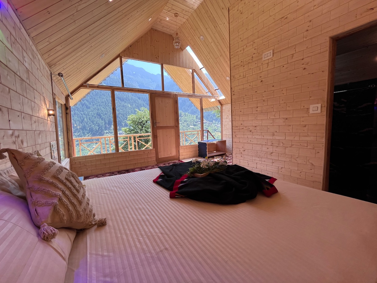 A bright bedroom features a spacious bed with neatly arranged linens. Large windows provide a view of the surrounding mountains, with natural light flooding the room. Wooden walls add warmth, while decorative elements enhance the cozy atmosphere.