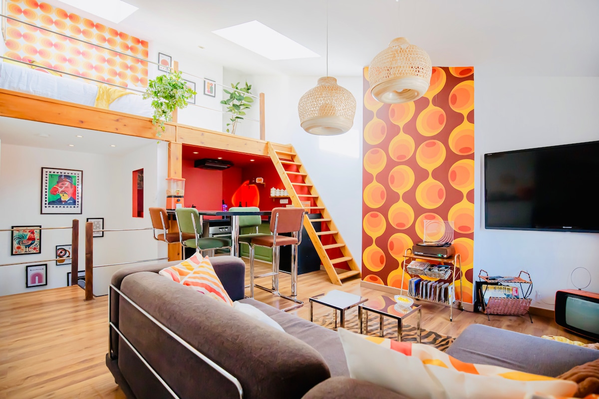 A bright open living space features a retro design with bold wallpaper and wooden accents. A cozy seating area with a large sofa is complemented by a TV unit displaying vintage decor. An elevated loft bed area is visible, highlighting the unique architecture.