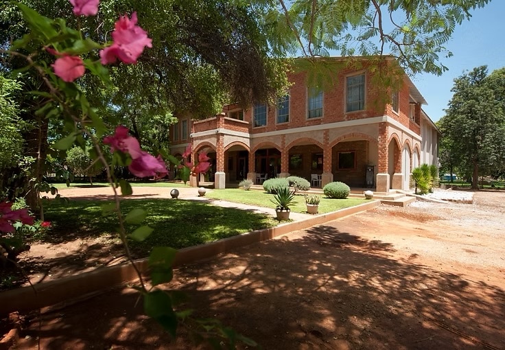 The exterior of the venue is showcased, featuring a two-story brick building with arched balconies and large windows. Lush greenery surrounds the property, complemented by flowering plants in the foreground. Well-maintained pathways lead to the entrance, creating a welcoming atmosphere.