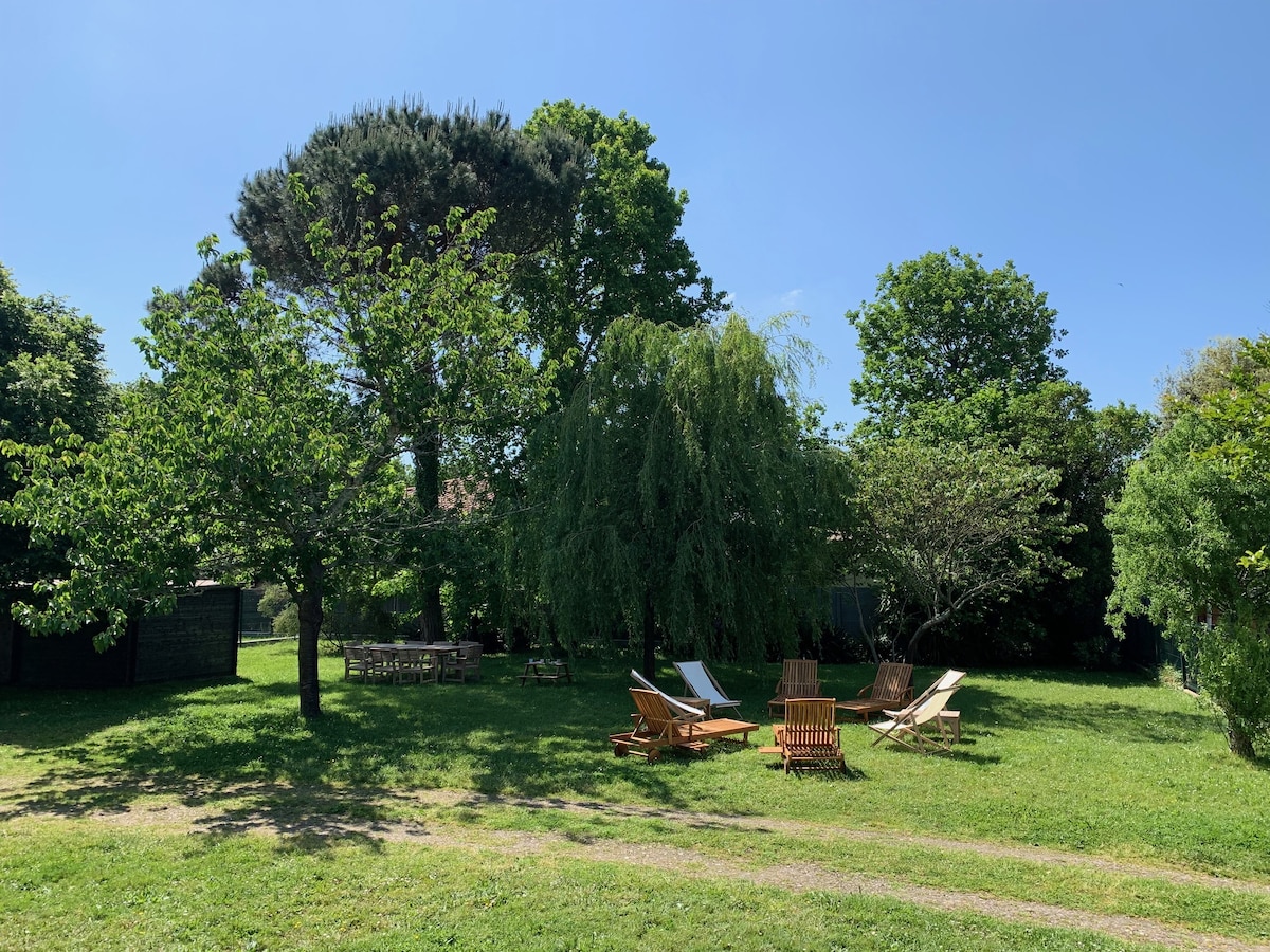 A spacious garden is featured, with various trees providing shade. Several wooden lounge chairs and sunbeds are arranged on the green grass, creating a suitable area for relaxation. Sunlight illuminates the space under a clear blue sky, while picnic tables are partially visible in the background.