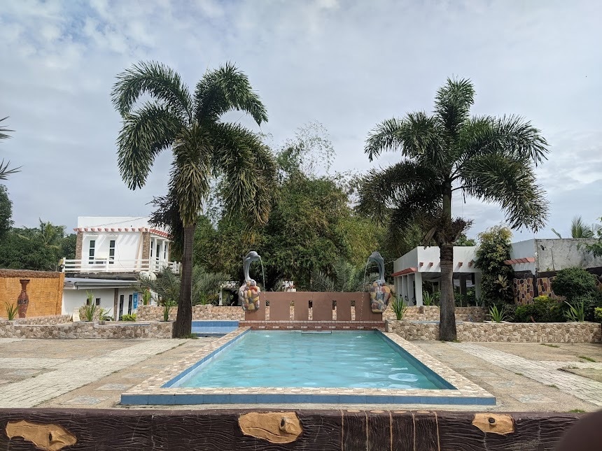 A clear pool is centrally positioned, bordered by textured stone paving and complemented by tall palm trees. The surrounding greenery adds to the serene environment, while a distinctive sculpture features at the head of the pool, framed by the resort's architecture in the background.