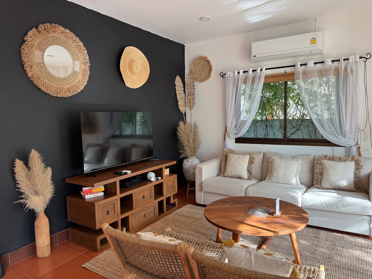 A living area is presented with a light-colored sectional sofa and a round wooden coffee table. A television is mounted on a dark accent wall, while decorative elements including mirrors and woven art enhance the space. Natural light filters through a window adorned with sheer curtains.