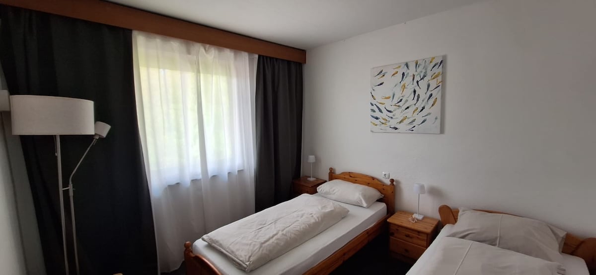 A bright bedroom is presented with two single beds featuring plain white linens. Natural light enters through a large window, complementing the soft color palette. A piece of artwork with abstract patterns adorns the wall above a bedside table, and a floor lamp adds additional lighting.