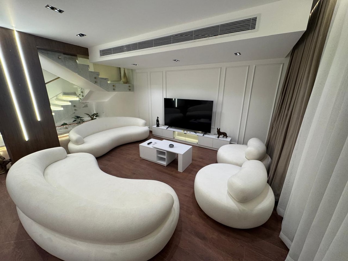 A modern living area is presented with curved, white seating arrangements and a minimalist coffee table. A large wall-mounted TV is positioned against a backdrop of soft textured panels, while warm wood flooring complements the overall design. Natural light streams through sheer curtains.