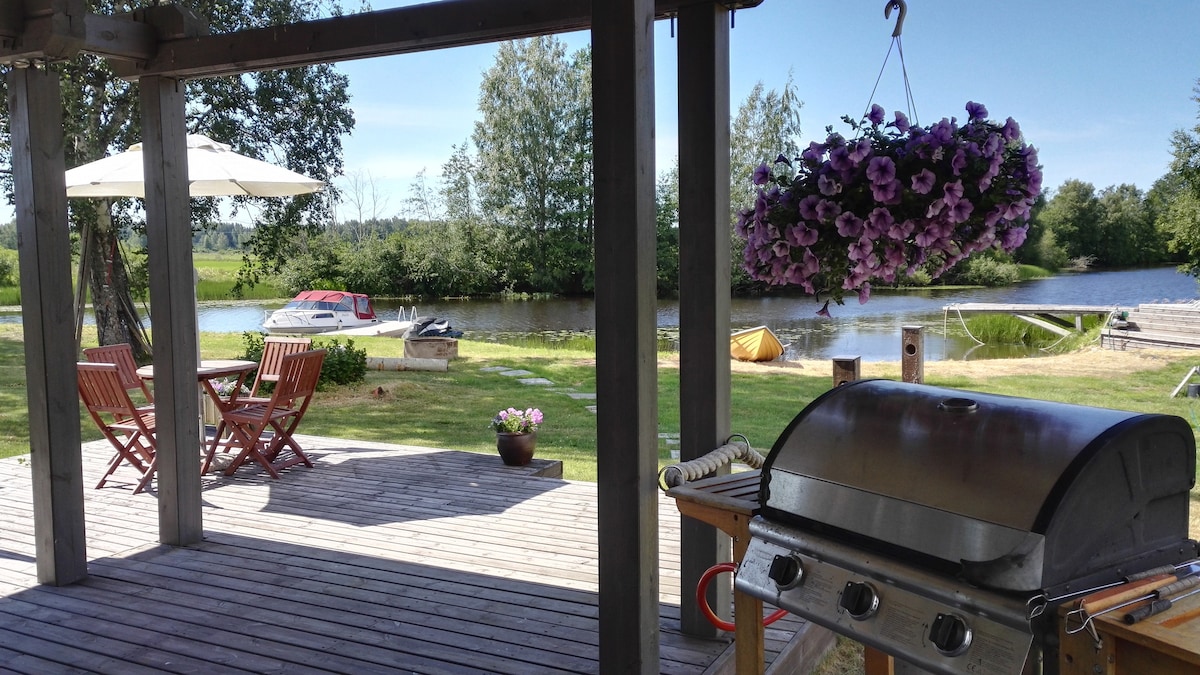 An outdoor terrace is featured, overlooking a calm river. A barbecue grill and a potted plant are visible nearby. A dining set with two chairs and a table are positioned under a shade umbrella. Colorful flowers hang overhead, and a canoe is seen in the distance.