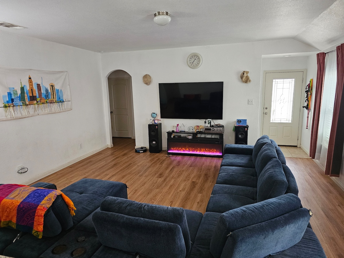 A comfortable living area is presented with a large, dark sectional sofa inviting relaxation. A flat-screen television is mounted on a white wall, with a colorful artwork displayed nearby. Natural light filters through a door leading outside, enhancing the spacious ambiance.