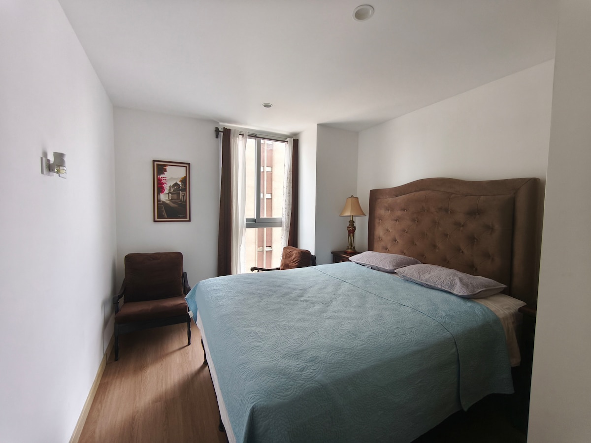 A cozy bedroom is illuminated by natural light from a large window. A spacious bed is adorned with a light blue coverlet. A comfortable armchair and a bedside table with a lamp provide additional seating and lighting options. Neutral tones create a relaxing environment.