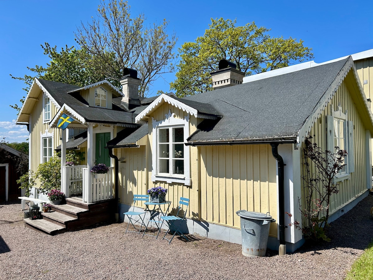 A charming yellow wooden house is presented, featuring a peaked roof and decorative trim. A small porch adorned with plants and two blue chairs invites relaxation. The background is complemented by greenery and a clear blue sky, adding to the overall serene setting.
