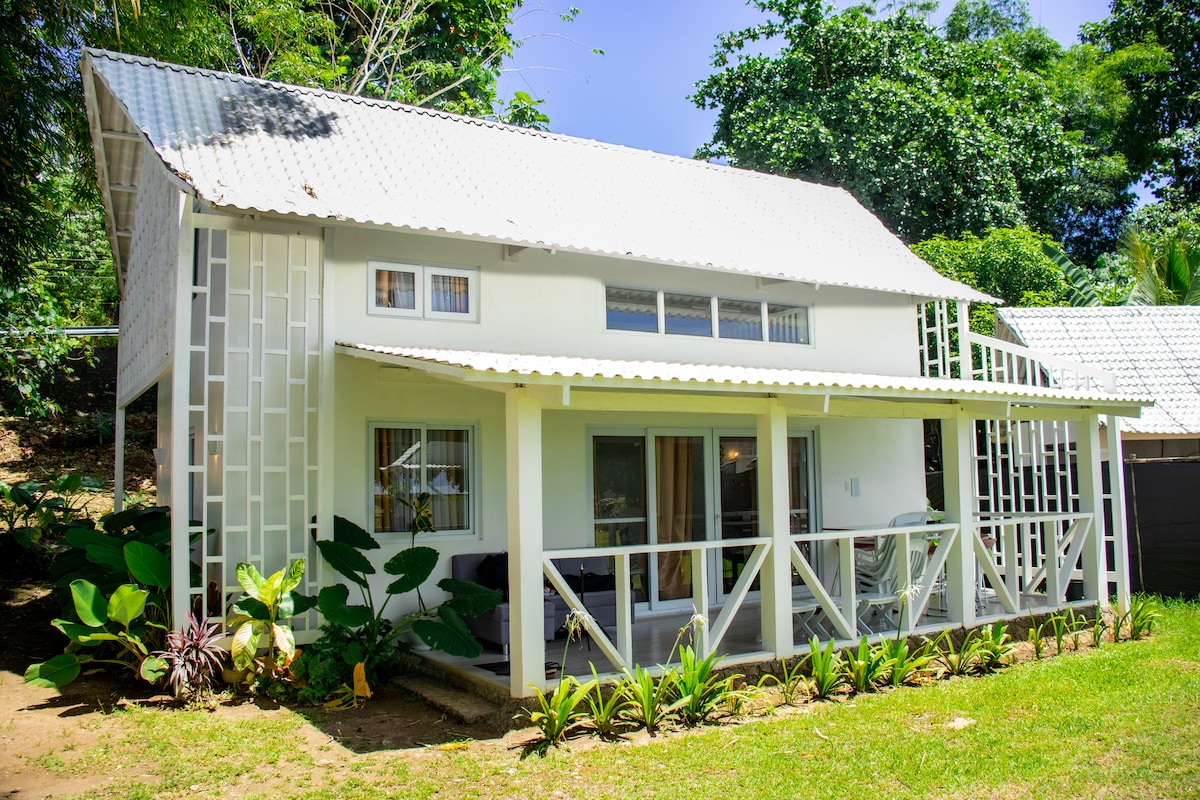 A two-storey villa features a light-colored exterior with a textured roof and large windows. Lush greenery surrounds the property, enhancing its natural setting. A covered porch with a railing invites relaxation while allowing views of the vibrant plants in the garden.