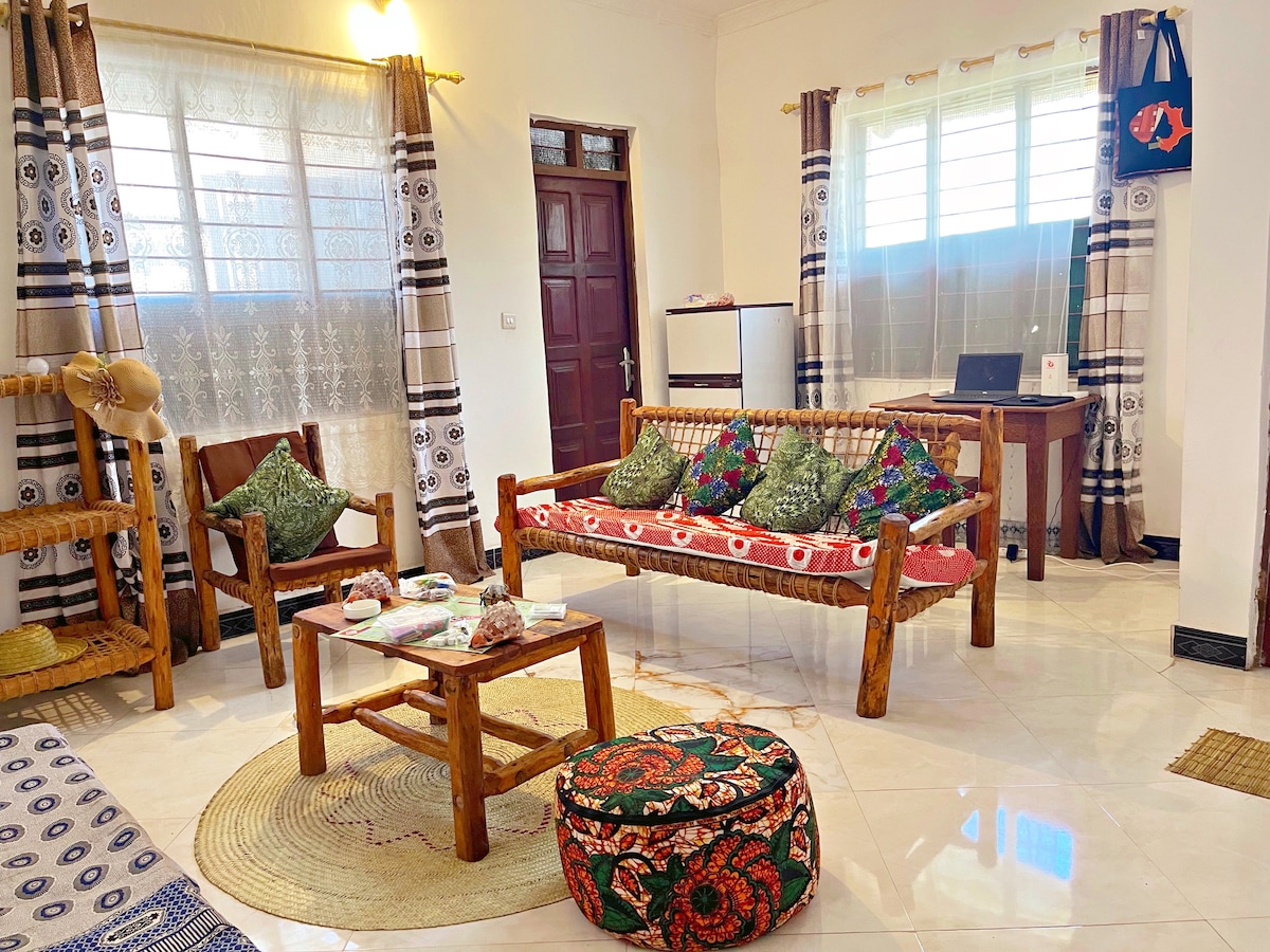 A comfortable living room area is showcased, featuring a wooden seating arrangement with colorful cushions. A round coffee table holds decorative items, while a patterned pouf adds a pop of color. Large windows allow natural light to fill the space, enhancing the inviting atmosphere.