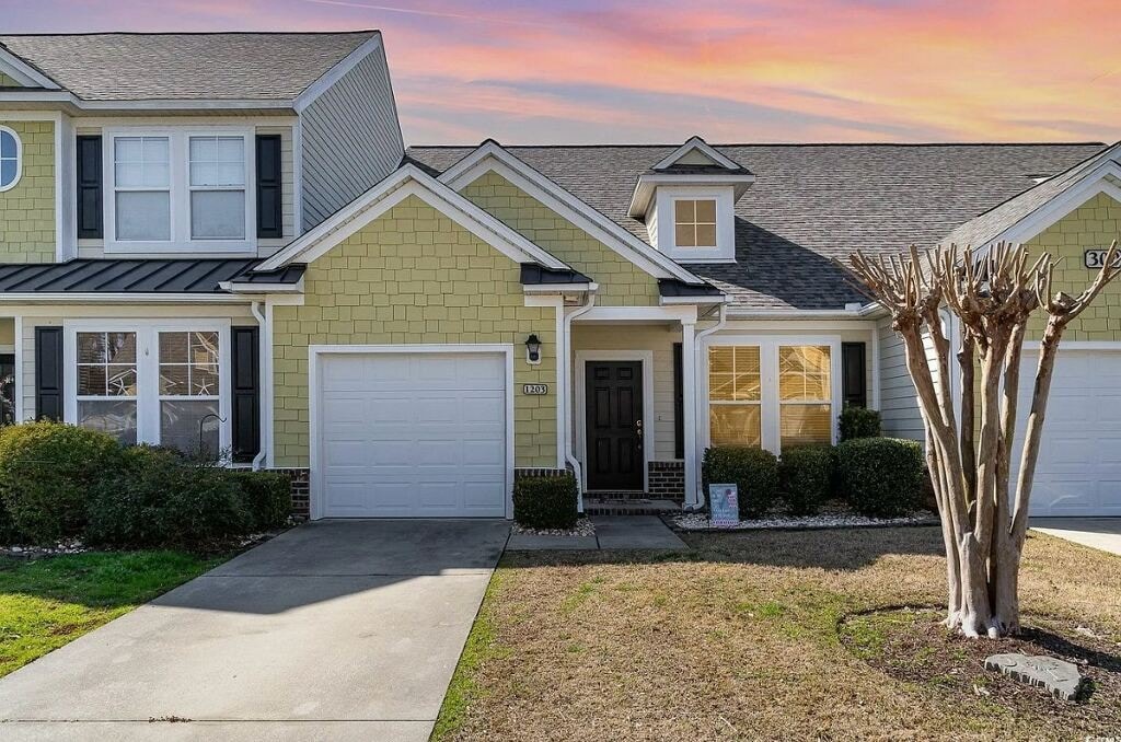 The exterior of a townhome showcases a charming combination of yellow siding and dark shutters. A two-car garage is visible on the left, and the entrance features a dark front door under a covered porch. Lush green landscaping complements the well-kept front yard.