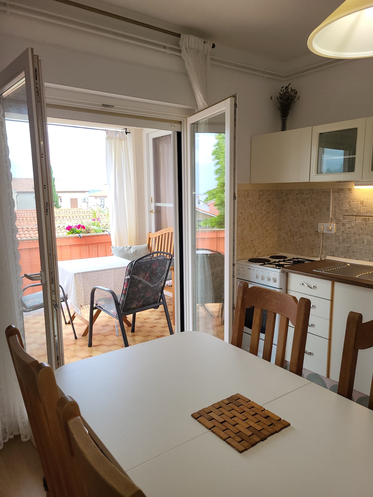 A kitchen area is visible with a dining table and wooden chairs. Glass doors lead to a balcony, where outdoor seating is arranged with a table and chairs. Natural light enters through the doors, and potted plants adorn the balcony.