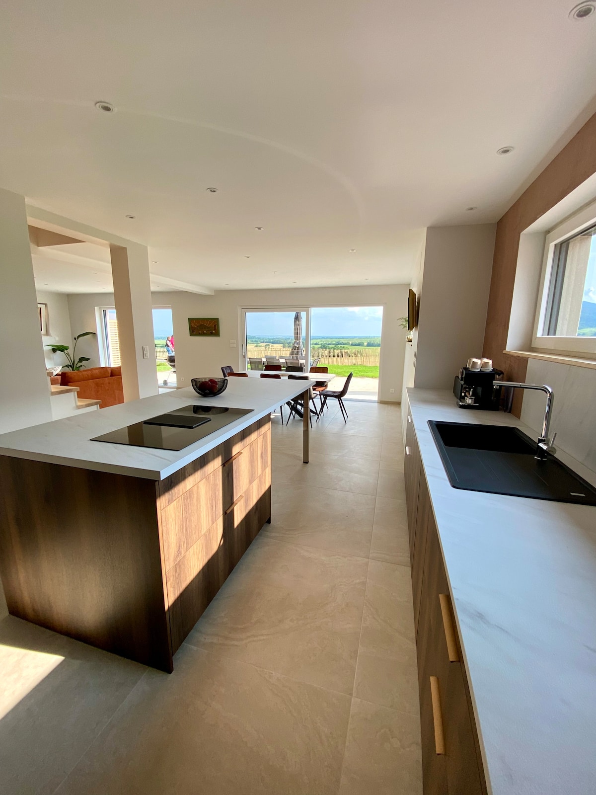 A spacious kitchen features a central island and modern appliances. The sleek countertops complement wooden cabinetry, creating a contemporary look. Large windows allow natural light to fill the space while offering views of the surrounding vineyard and countryside.