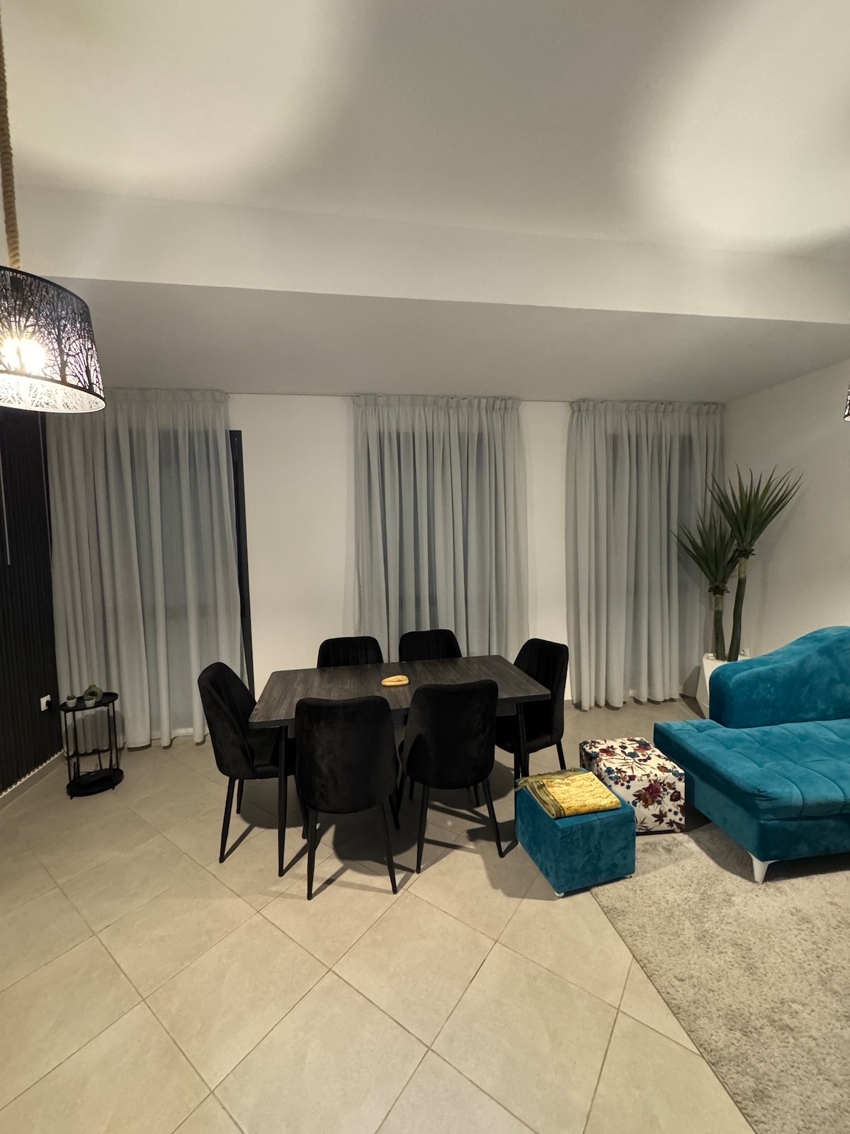 A dining area is showcased with a dark table surrounded by six plush chairs. Two large windows, dressed in sheer curtains, allow soft light to fill the space. A cozy seating arrangement with a turquoise sofa and a decorative ottoman is visible in the corner.