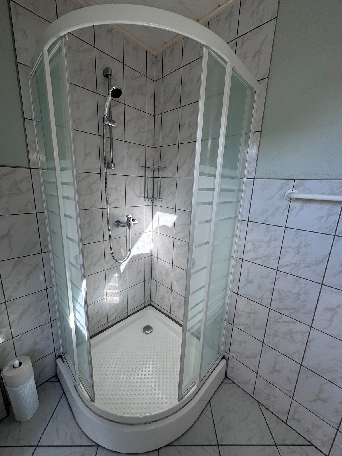 A shower enclosure features clear glass doors, allowing natural light to enter. White tiled walls and a textured floor provide a clean and functional design. Shower fixtures are mounted on the wall, with a small shelf adjacent to them for toiletries.