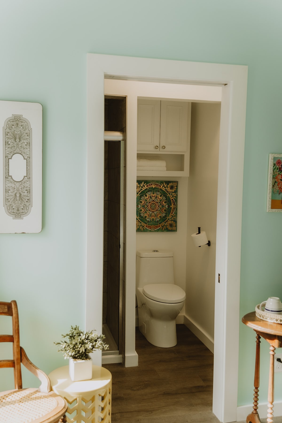 A compact bathroom area is visible through an open door. It features a toilet and a small shower space. Colorful wall art adds a decorative touch to the room, while light teal walls create a fresh ambiance. A wooden chair and a small table with a potted plant occupy the adjacent space.