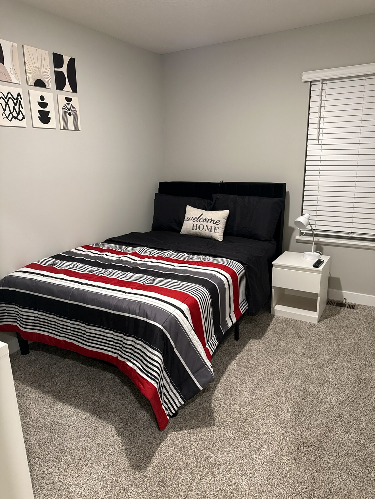 A cozy bedroom is presented with a black headboard and a striped bedspread in shades of gray, red, and white. A small bedside table with a lamp is positioned on one side, while a framed wall art set hangs above the bed. Soft carpeting is visible on the floor.
