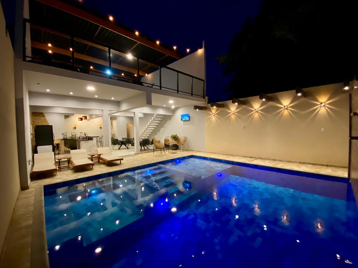 An illuminated outdoor area features a heated pool with blue water reflecting lights. Surrounding the pool, lounge chairs are positioned for relaxation. A second-level terrace overlooks the space, while a softly lit wall defines the perimeter, creating a serene atmosphere during nighttime.