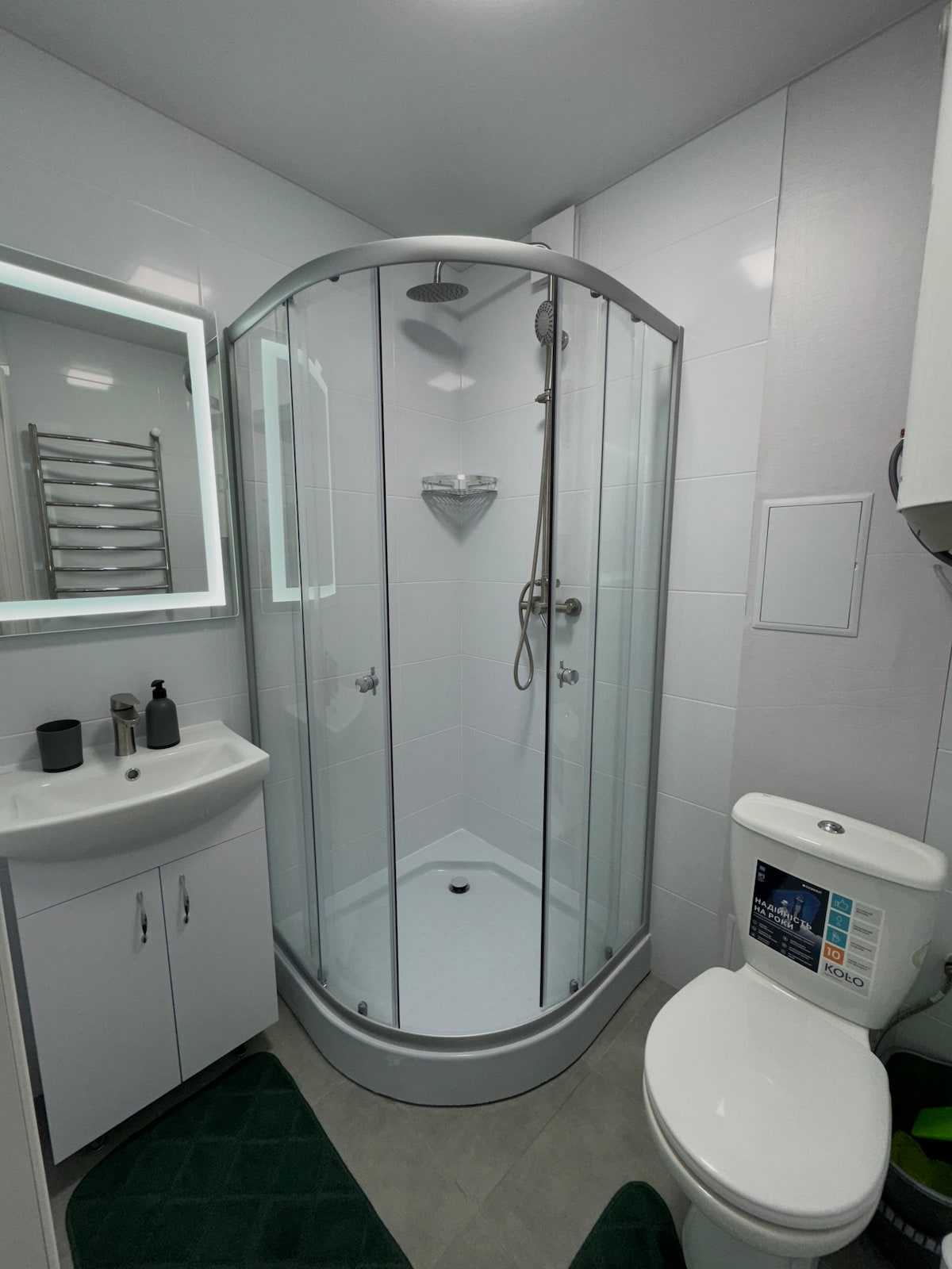 A modern bathroom features a glass-enclosed shower with a handheld showerhead and wall-mounted fixtures. A sink with a minimalist design is accompanied by grooming products. A towel warmer is mounted on the wall, and a toilet is positioned adjacent to the shower. Neutral tiles complete the space.