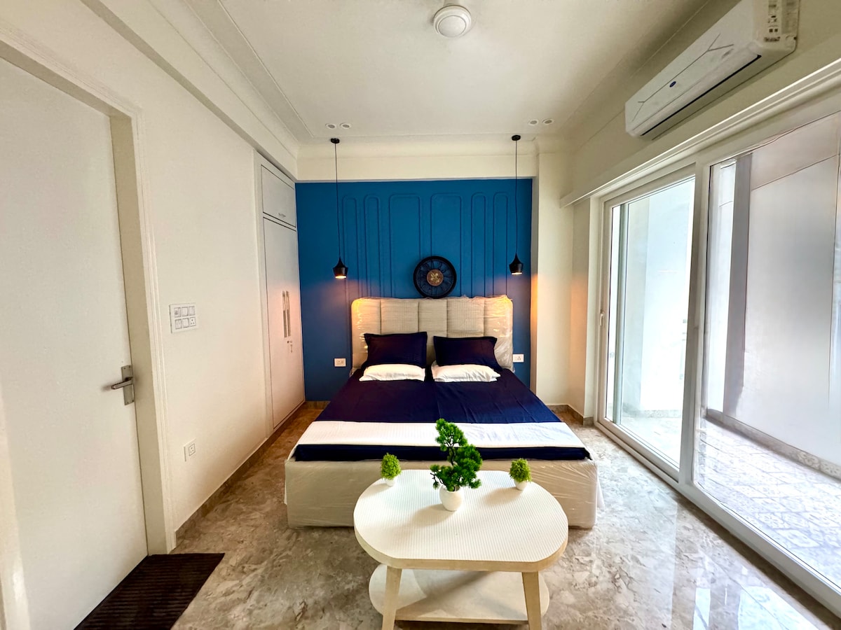 A bright bedroom features a spacious bed with dark bedding against a bold blue accent wall. Natural light enters through large glass doors, illuminating a small round table with a potted plant. Air conditioning units are visible on the wall, adding comfort.