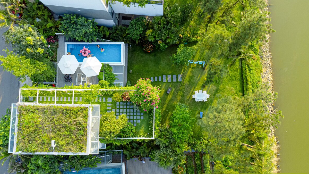 An aerial view reveals a serene villa complex, featuring manicured gardens and a private swimming pool. Lounge chairs under umbrellas are surrounded by lush greenery. A pathway lined with stones leads to a dining area set in the garden, adjacent to the water's edge.
