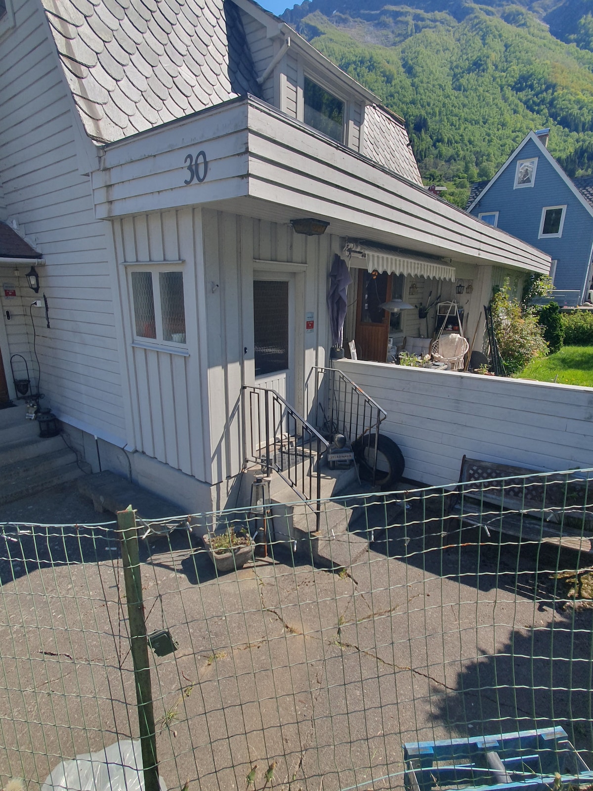 An inviting entrance of a light-colored house is framed by a small set of steps leading to a door. A porch area is visible, adorned with outdoor furnishings. A fenced yard, with elements of greenery and a paved pathway, completes the view of this charming home.