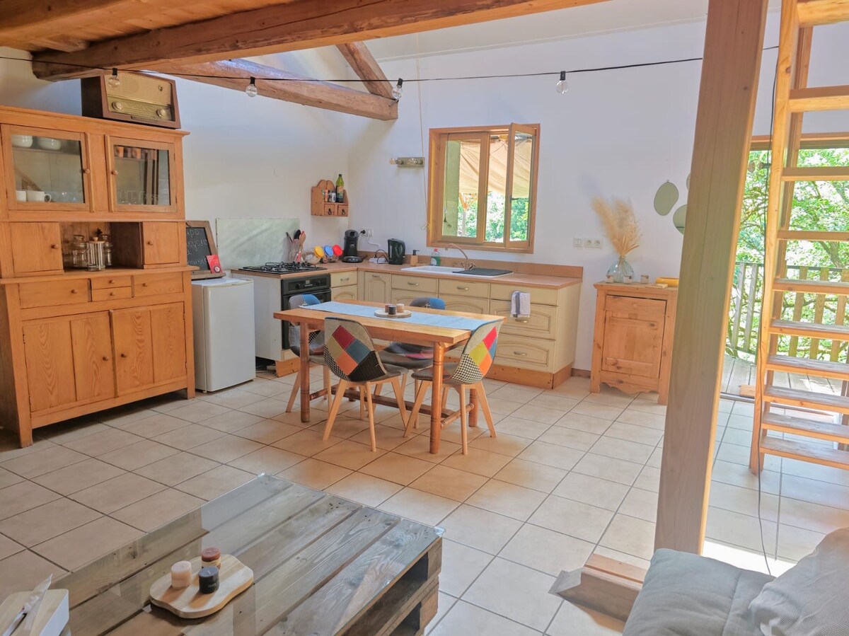 A spacious kitchen area is highlighted by wooden beams and natural light streaming through a window. A wooden table with colorful chairs sits at the center, surrounded by cabinetry. The space includes a stove and sink, contributing to a cozy cooking environment.