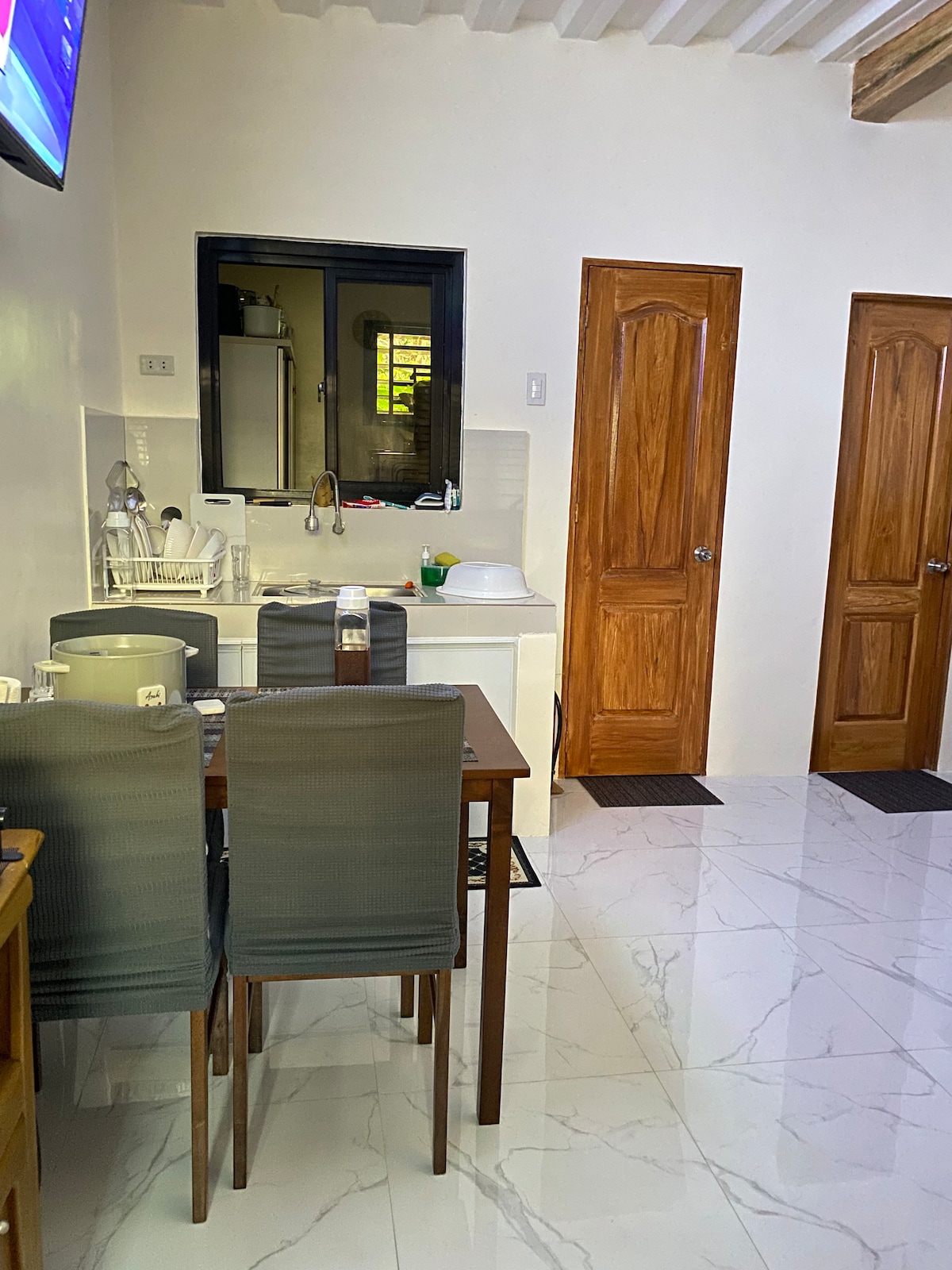 A bright kitchen area is visible, featuring a dining table set for four. Modern cabinetry is complemented by a sleek sink and countertop. Two wooden doors lead to separate rooms, while a television mounted in the corner adds convenience.
