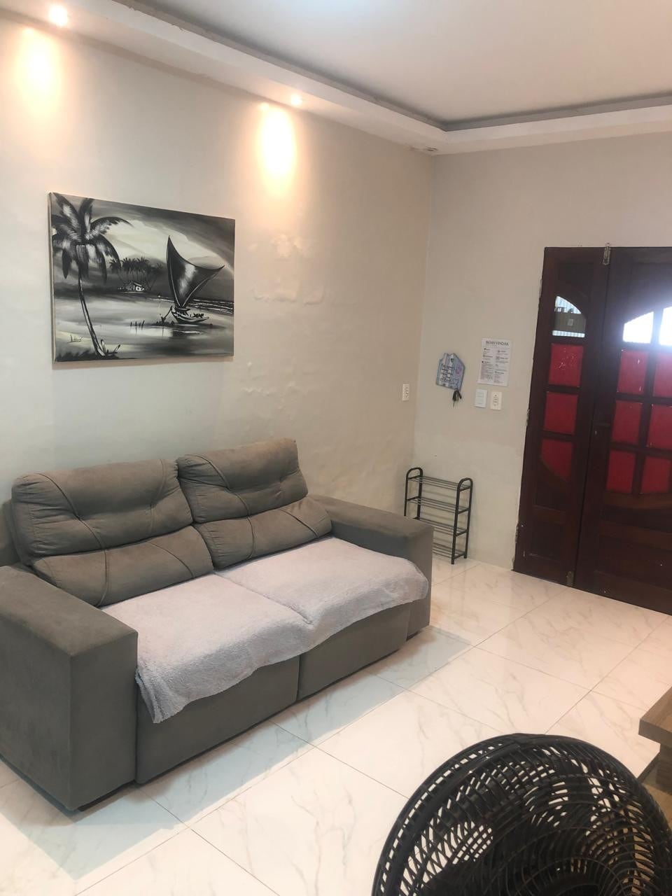 A cozy sitting area is featured with a gray sofa, complemented by a light throw blanket. A black and white artwork depicting a sailboat is hung above the sofa. Natural light is provided by the entrance door, which has traditional wooden detailing.