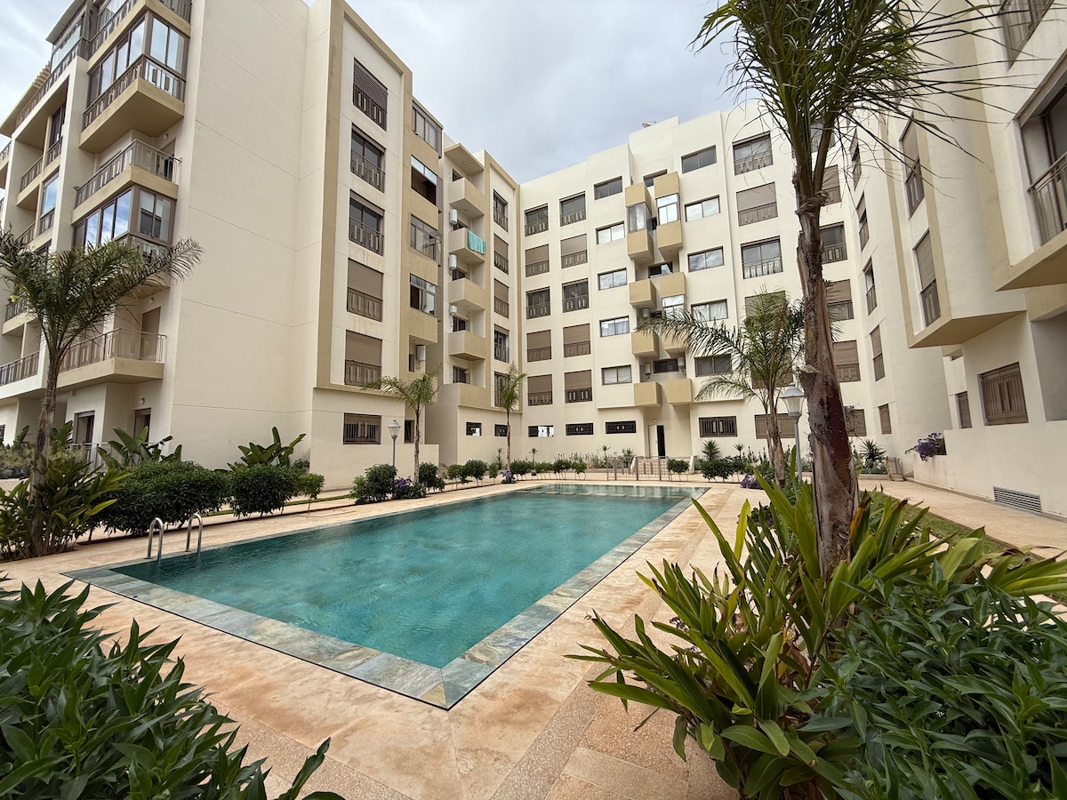 A refreshing outdoor pool is framed by landscaped greenery, with palm trees and shrubs surrounding the area. The modern building features multiple floors with large windows, creating an airy environment. A seating area is positioned near the water, offering space for relaxation.