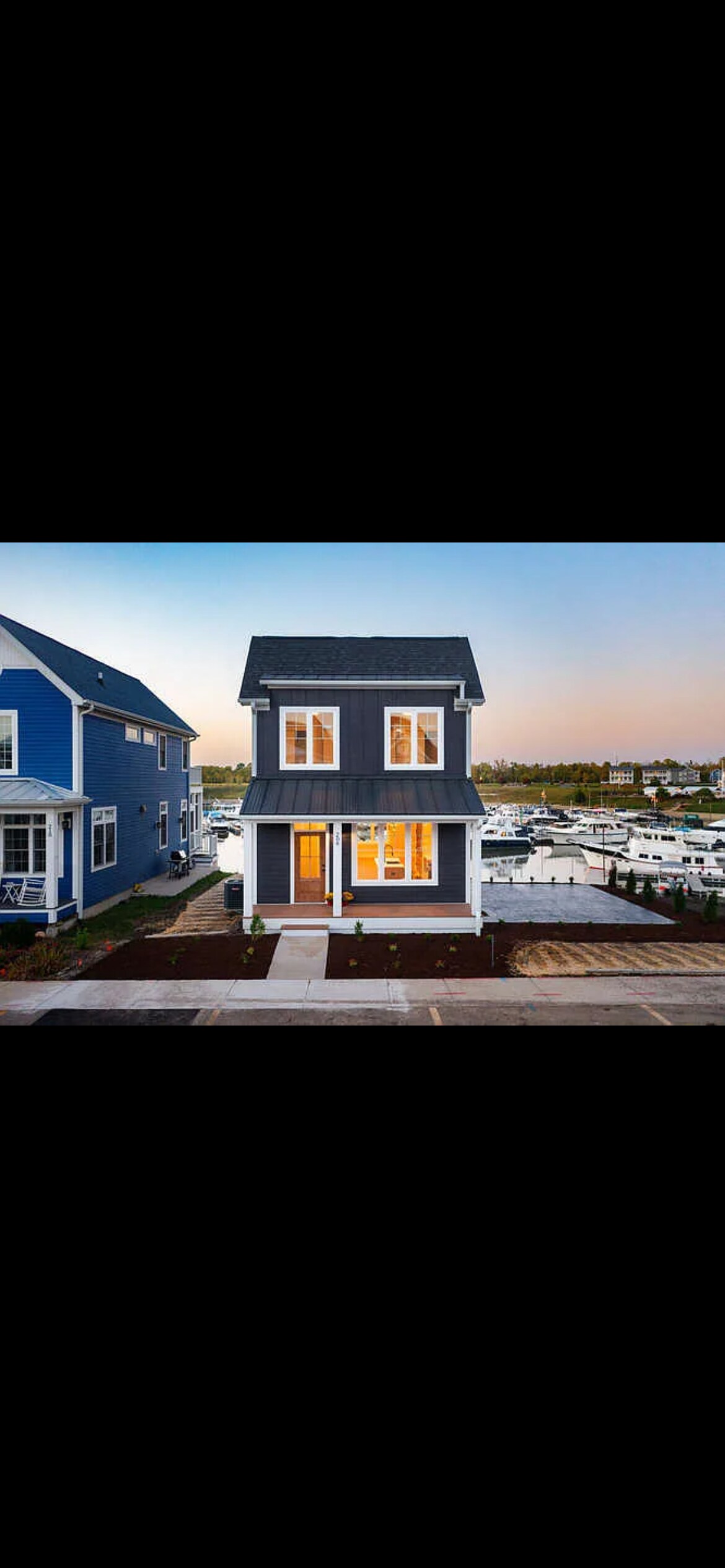 A three-story home with a blue exterior is positioned near a marina. Large windows are present on each floor, allowing ample natural light. The front door is illuminated, creating a welcoming entrance. Nearby, well-maintained landscaping complements the structure.