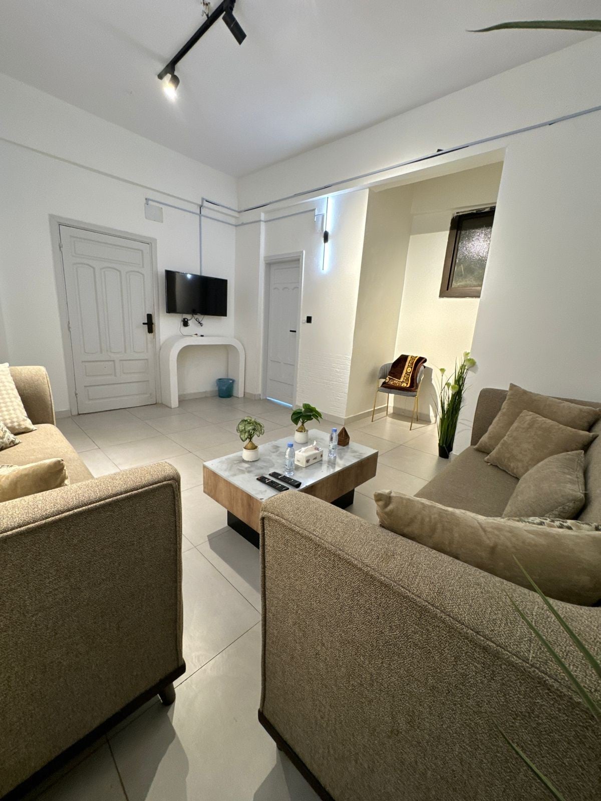 A light-filled living area features two beige sofas positioned around a wooden coffee table. A wall-mounted TV is visible, along with a subtle decorative piece near a doorway. The space is complemented by indoor plants, enhancing the overall ambiance.